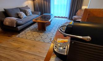 A cozy living space with a gray sofa, a wooden table, and a coffee machine. The window lets in a lot of daylight and the room is tastefully decorated.