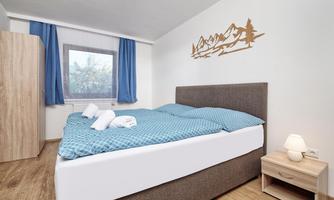 A modern bedroom with a large bed and blue pillows. The wall features carved wooden mountains, and the room has large windows with curtains.