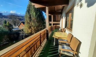 An inviting balcony with cozy chairs and a table. In the background, trees and a clear sky are visible.