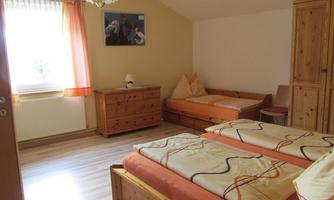 A cozy bedroom with two beds and wooden furniture. The room has large windows with bright curtains and a warm, inviting ambiance.