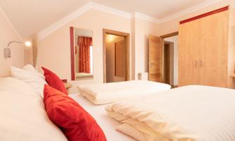 A bright bedroom with two beds, red pillows, and a wooden wardrobe. The walls are painted in soft colors, and there are windows with curtains.