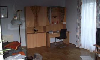 A simply furnished room with a desk and a chair. There is a couch and large windows with curtains.