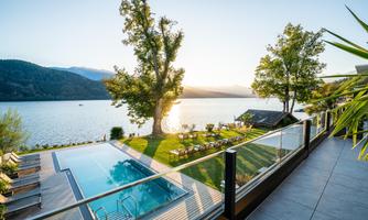 A beautiful view of a lake with an infinity pool and loungers. The sun sets behind the mountains, illuminating the surroundings.