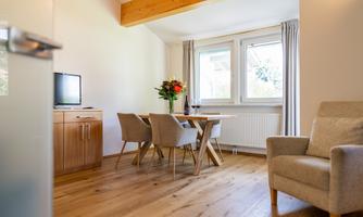 A bright living area with a dining table and modern chairs. A cozy armchair and a vase of flowers create an inviting atmosphere.