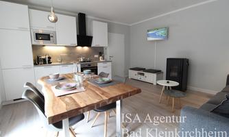 A modern kitchen with a large wooden dining table and elegant chairs. In the background, a living room and a small seating area with a television are visible.
