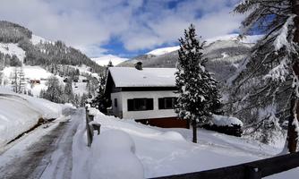A charming house in the snow with snow-covered trees and mountains in the background. The sky is partly cloudy, giving the scene a wintry atmosphere.