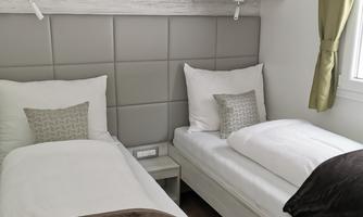 A bright room with two single beds and modern furniture. The walls are light grey and a window with green curtains lets in daylight.