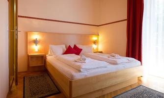A cozy bedroom with a double bed and red pillows. The room has large windows and warm light.