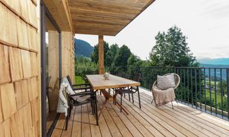 A modern wooden terrace with a large table and chairs. In the background, green trees and mountains can be seen.