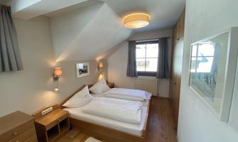 A cozy bedroom with two beds, a wooden table, and a window with curtains. The walls are bright, and the floor is covered with wood.