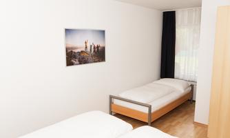 A simple bedroom with two single beds and a large window. On the wall hangs a picture of people in nature.