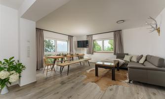 A modern living room with large windows and plenty of natural light. The furnishings include a gray sofa, a wooden dining table, and decorative plants.