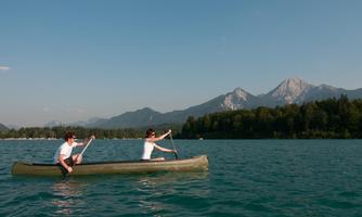 Two people are paddling in a canoe on a clear lake. In the background, there are mountains and a blue sky.