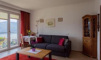 A cozy living room with a gray sofa, a wooden table, and a large window. The decor is simple, with red curtains and a red carpet.