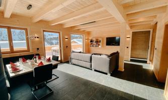 A cozy living room with wooden decor and large windows. In the foreground, there is a dining table, and the room is bright and inviting.