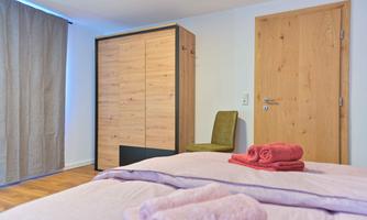 A bright bedroom with a wooden wardrobe and a chair. Pink towels are on the bed.