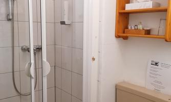 A small bathroom with a shower and a toilet. On the wall, there is a wooden shelf with toiletries.
