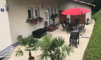 A cozy outdoor area with patio furniture and a red umbrella. Surrounded by plants and blooming windows.