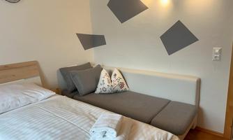 A simple bedroom with a single bed and a cozy sitting area. The walls are decorated with geometric patterns.