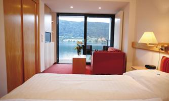 A modern room with a double bed and a view of the lake. In the background, there is a cozy living area with a sofa and a large window front.