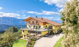 A picturesque country house with a yellow facade and large windows in a hilly landscape. In the background, there are mountains and a clear blue sky.