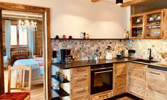 A modern kitchen with wooden furniture and colorful tiles. In the background, a cozy bedroom is visible.
