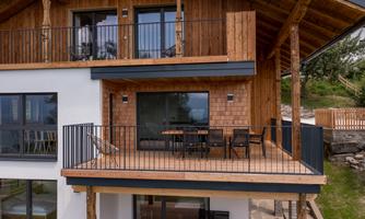 A modern wooden house with a large balcony and seating. The surroundings are close to nature and inviting.