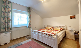 A cozy bedroom with a large white bed and colorful bedding. The walls are light, and there are curtains with a floral pattern.