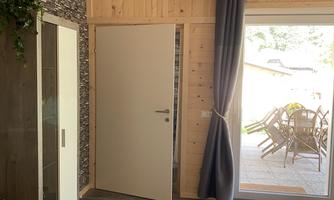 A bright room with wooden paneling, a TV on the wall, and an open door leading to an outdoor area. A cozy sofa and a plant box are also visible.