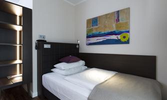 A modern bedroom with a single bed and a colorful mural. The room is bright and minimally furnished, with an open shelf in the background.
