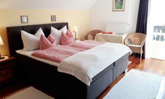 A cozy bedroom with a large double bed and pink cushions. The room has a light wooden floor and access to natural light.