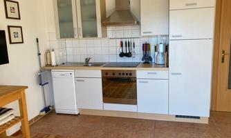A modern kitchen with white furniture and wooden flooring. It features a stove, a sink, and various kitchen appliances.