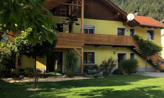 A beautiful house with wooden elements and a green garden. In the background, gentle hills and a blue sky can be seen.