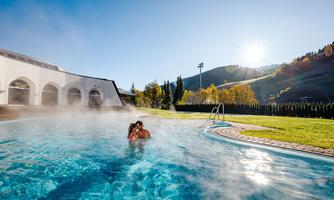 A romantic couple in the warm water of a pool, surrounded by a misty atmosphere. In the background, gentle hills rise and a clear blue sky.