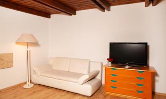 A cozy living room with a white sofa, a floor lamp, and a television. The floor is made of wood, and there is a colorful dresser.