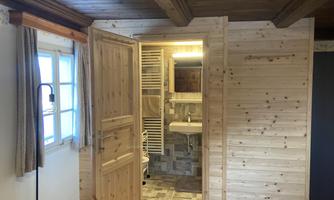 A natural room with wooden paneling and a small bathroom in the background. The room has a cozy atmosphere with warm light.