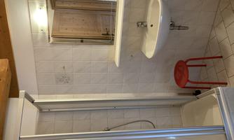 A small bathroom with a shower, a sink, and a red stool. The walls are tiled white and the floor is light.