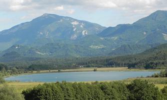 An idyllic landscape with a clear lake surrounded by mountains. In the foreground, there are green trees and meadows.