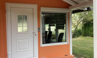 An orange holiday home with a white door and a large window. The outdoor area has gray tiles and a well-kept lawn.