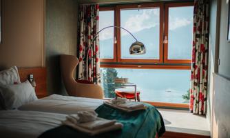A modern hotel room with two beds and a large window. The view shows a picturesque landscape with water and mountains in the background.