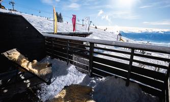 A balcony in the snow with deck chairs and a view of the slope. Behind the balcony, there are snowdrifts and a sunny mountain landscape visible.