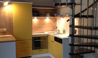 A modern kitchen with yellow cabinets and wooden details. An elegant spiral staircase leads to an upper area.