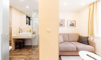 A modern apartment with a bright living area and a comfortable sofa. In the background, there is a bathroom with a glass sliding door.