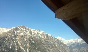 A majestic mountain landscape with snow-covered peaks under a clear blue sky. In the foreground, part of a wooden structure is visible.
