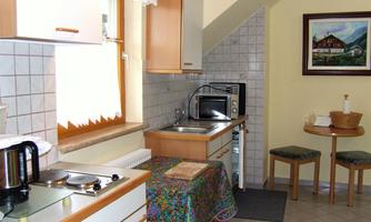 A small, cozy kitchen with wooden furniture and a table for two people. Visible appliances include a stove, a microwave, and a coffee maker.