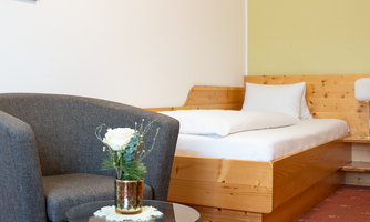 A cozy room with a wooden bed and a comfortable armchair. On a small table, there is a vase with a white flower.
