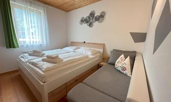 A cozy bedroom with a double bed and an additional sofa. The walls are plain, and a window brings daylight into the room.