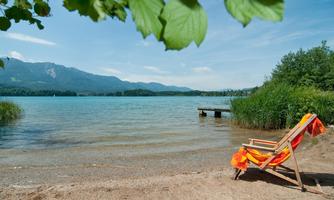 A tranquil lake with clear water and gentle waves. A deck chair with a colorful towel is by the shore, surrounded by green plants and mountains in the background.