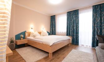 A cozy bedroom with a large bed and light curtains. The room has wooden floors and modern decoration.