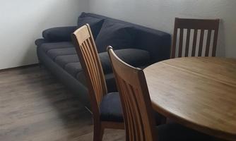 A modern living space with a gray sofa and a round wooden table. There are four wooden chairs and plenty of daylight streaming through the curtains.
